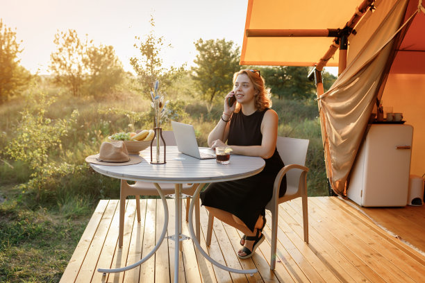 在一个阳光明媚的日子里，在舒适的豪华露营帐篷里工作时，微笑的自由职业者正在打电话。豪华户外露营帐篷，适合夏季度假度假。生活方式的概念图片下载