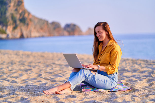 年轻快乐快乐的微笑无忧无虑满意的千禧一代自由职业者妇女使用电脑和坐在沙滩上的海边。梦想办公室远程办公概念图片下载