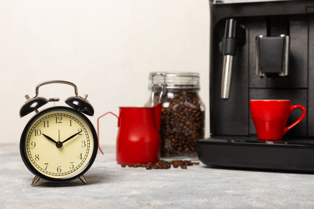 咖啡机的背景是闹钟。黑色闹钟放在厨房的桌子上，背景是一杯芳香的咖啡和一台咖啡机。愉快的早晨概念。图片下载