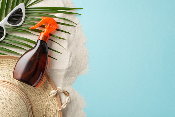 在阳光下保持安全。无品牌防晒油瓶俯视图，配以淡蓝色和沙质背景的太阳镜、帽子、贝壳手镯、棕榈叶，为标签或广告提供空白空间图片下载