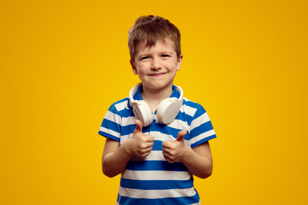 可爱的孩子穿着蓝色条纹polo衫，脖子上戴着耳机，大拇指朝上，孤立在黄色背景下图片下载
