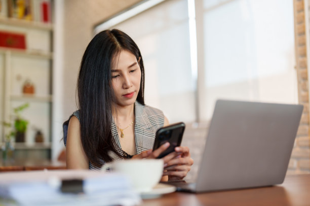 特写，漂亮的亚洲女人坐在手机上，微笑着通过手机上的聊天应用和男朋友聊天，桌子上有笔记本电脑，这是女人工作用的电脑。图片下载
