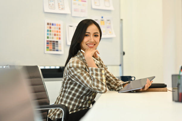 自信的女性web开发人员手持数字平板电脑，坐在创意办公室，对着镜头微笑。图片下载