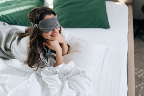 在酒店豪华的卧室里，微笑的年轻女子戴着睡眠面罩躺在舒适的床上。穿着睡衣的黑发美女肖像图片下载