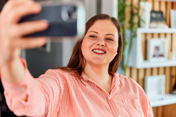 一个穿着漂亮条纹衬衫、微笑着、曲线优美的女人拿着手机自拍。图片下载