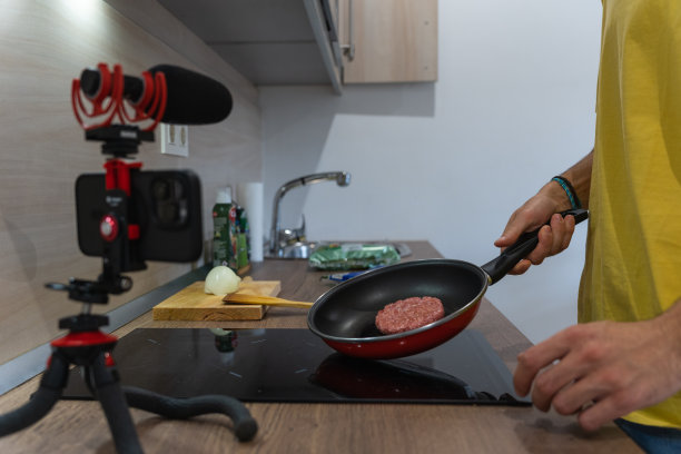 一名无法辨认的男子在家里的现代厨房里煮汉堡，同时用智能手机和麦克风录下自己的声音。一名男子在平底锅上准备肉，并将其录制在视频博客或社交媒体上。图片下载