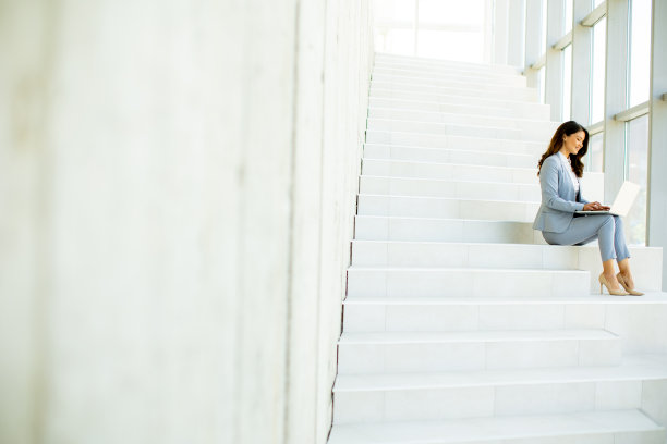 年轻的女商人坐在办公室走廊的楼梯上，用笔记本电脑工作图片下载