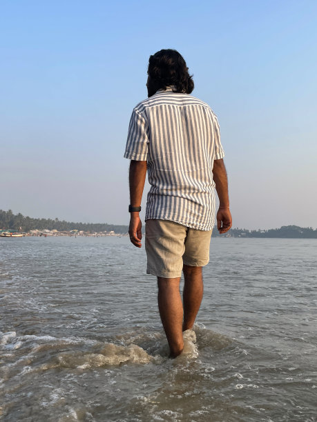 印度果阿帕洛莱姆，沙滩上穿着短裤和条纹短袖衬衫的印度男子的特写照片，黄昏时，度假游客光着脚走在海边，后景，重点在前景图片下载