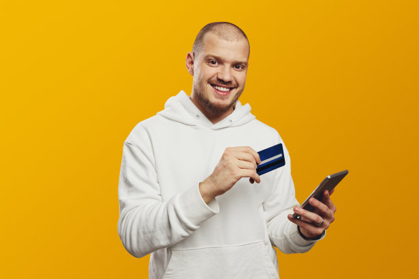 身穿白色连帽衫、留着胡子的年轻男子拿着信用卡和智能手机，孤立地出现在黄色背景中图片下载