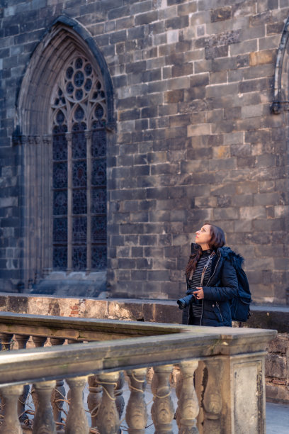 在西班牙观光旅行中，体贴的年轻女子拿着相机站在砖墙上欣赏哥特区古老的巴塞罗那大教堂图片下载