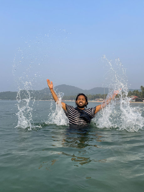 印度果阿帕洛莱姆海滩，一名印度男子在海滩度假，手臂举起，穿着黑白相间的t恤，胸部深埋在海水中，胸前系着防水袋，波涛汹涌的水面上，印度果阿帕洛莱姆海滩图片下载