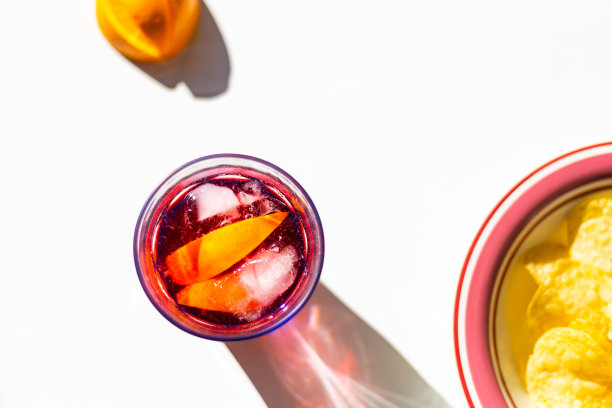 俯视图，在阳光明媚的白色餐桌上，玻璃杯里放着红色烈酒(马提尼、苦艾酒、桑格利亚酒、苦酒)、橙子和冰块，旁边是一盘薯片图片下载