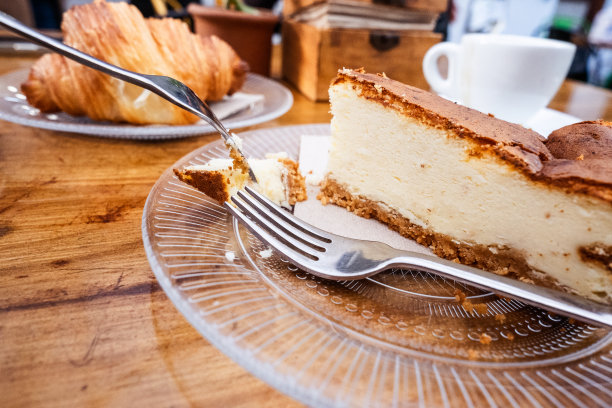早餐。一块芝士蛋糕放在玻璃板上用金属叉子，桌子上有一杯咖啡放在盘子上用金属勺子，玻璃板上放着一个牛角面包。一切都在一张天然木桌上。美食。西班牙加那利群岛的特内里费岛图片下载