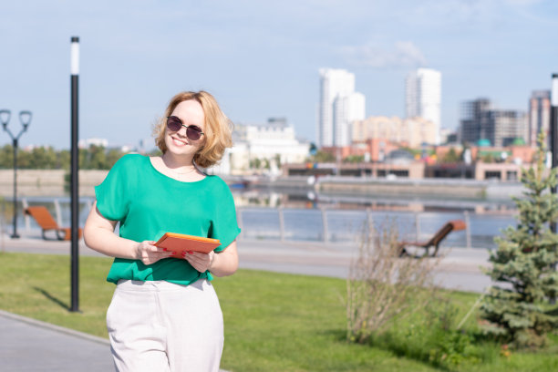 一个面带微笑的女学生，戴着墨镜，手里拿着笔记本或平板电脑，站在河堤上，在街上。图片下载