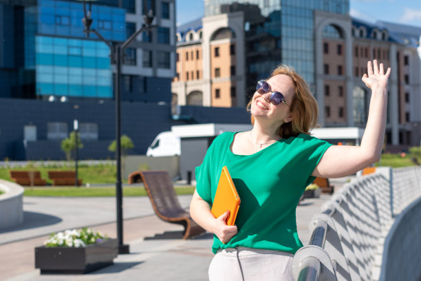 一个戴着墨镜微笑的女学生，拿着数码平板电脑，在街上挥手和朋友打招呼。图片下载