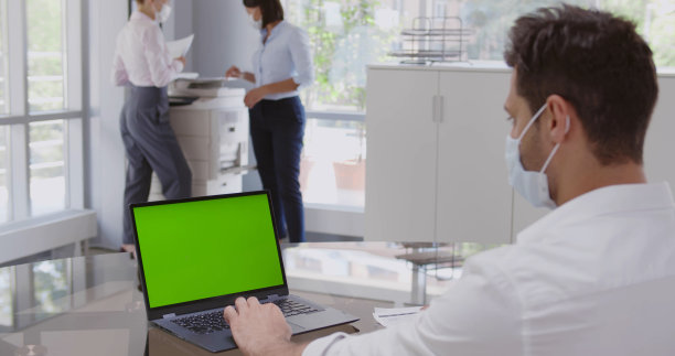 回头看戴着防护面具的年轻成功男子坐在办公室的办公桌前，在电脑前工作。图片下载