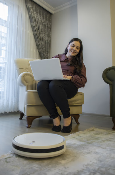 一个女人在家里用电脑工作，一个机器人吸尘器在打扫房子图片下载