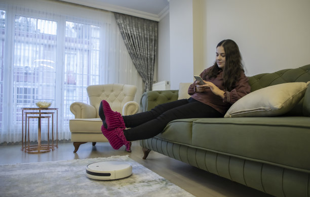 一个女人在家里用电话工作，一个机器人吸尘器在打扫房子图片下载