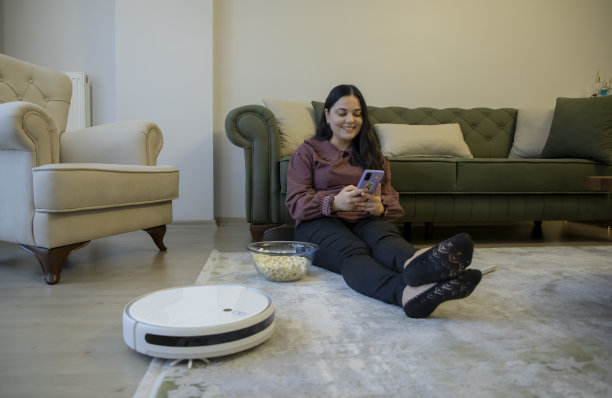 一个女人在家里用电话工作，一个机器人吸尘器在打扫房子图片下载