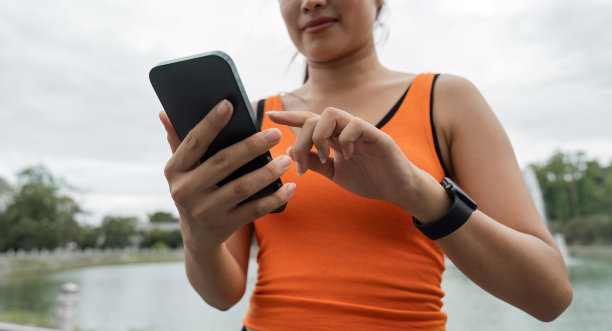 微笑的亚洲女性的肖像，看着她的智能手机和阅读应用程序，穿着运动服站在公园里图片下载