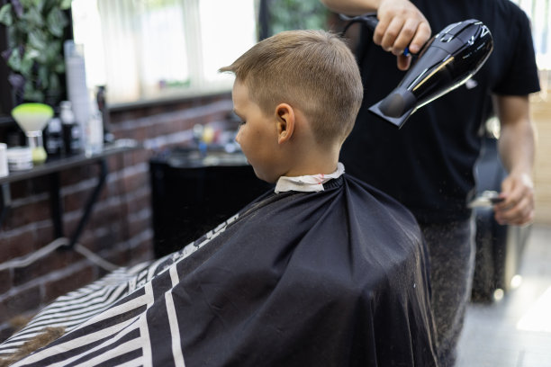 在吹风机的帮助下，公园管理员吹掉了男孩孩子脖子和肩膀上剪下来的头发。理发师为这个夏天的孩子做了一个时髦的发型。图片下载