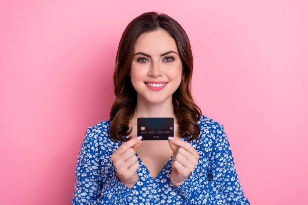 照片乐观可爱可爱的女人波浪发型穿蓝色连衣裙手拿信用卡塑料卡孤立在粉红色的背景图片下载