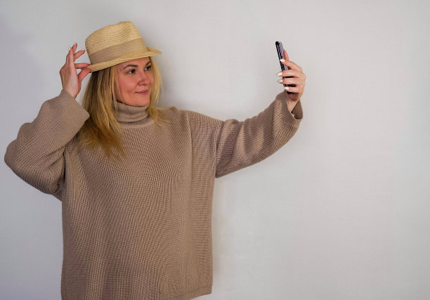 一个金发女孩，头戴草帽，身穿棕色毛衣，站在白色的墙上自拍图片下载