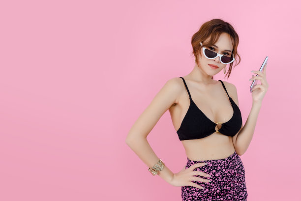 性感美女穿着内衣比基尼，太阳镜拿着智能手机粉红色的背景。写真美女用手机。暑假概念。图片下载