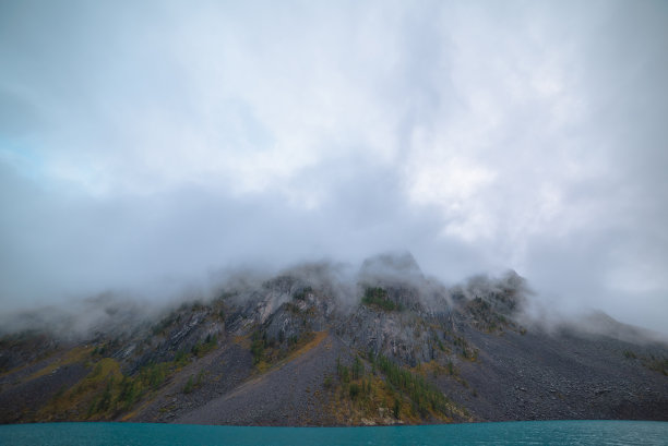 戏剧性的宁静冥想景观与涟漪在大绿松石山湖对高尖锐的岩石在低云。平静蔚蓝的高山湖泊和巨大的岩石山脉在低多云灰色的天空。图片下载