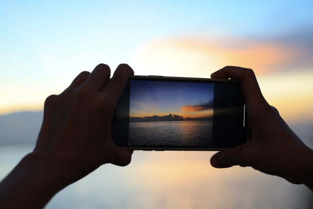 一个女人拿着手机拍摄明亮的日落和大海的特写镜头。手持智能手机拍摄日落时的大海图片下载