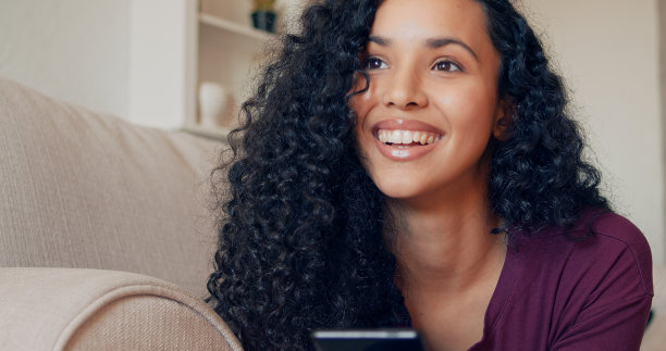 开朗的非洲年轻女子坐在沙发上玩智能手机。黑人微笑女子在家里用手机上的应用程序。漂亮的女孩一边聊天一边放松。图片下载