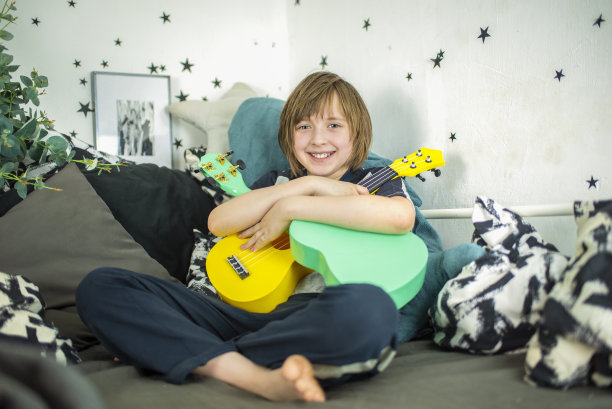 一个小男孩抱着一把黄色的尤克里里琴，一个快乐的孩子坐在他的床上，在黑白相间的床上，弹着迷你吉他，唱着歌图片下载