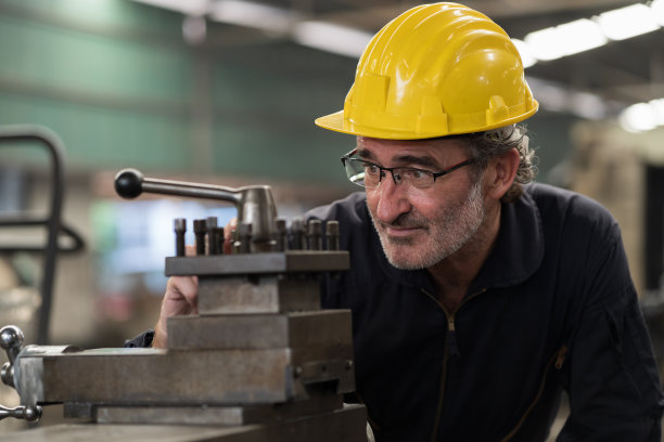 在工业工厂检查车床零件质量的高级男工程师，穿着安全制服，头戴安全帽。成熟的男性技术工人维护机器零件图片下载