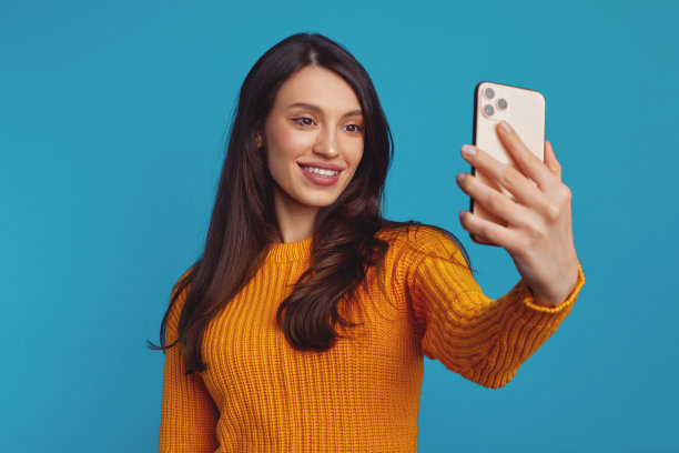 穿着橙色毛衣的可爱女孩拿着智能手机自拍，被蓝色背景隔离图片下载