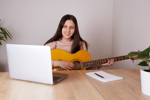 一个快乐的女孩正在学习弹吉他。一个青少年正坐在笔记本电脑前的桌子上观看在线课程。图片下载