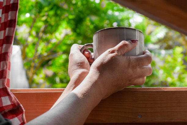 从一个女人手里端着一杯咖啡，站在窗外俯瞰外面或花园的早晨景色。图片下载