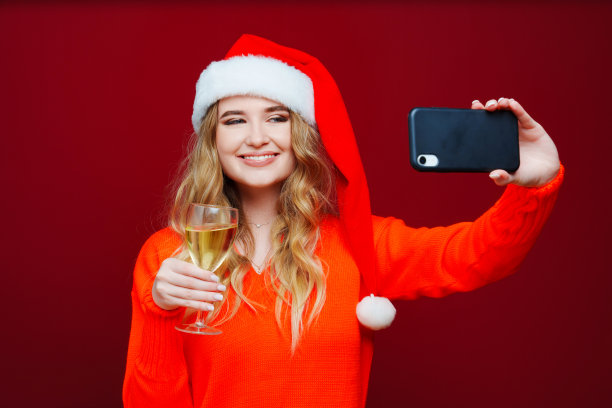 一个女人戴着圣诞老人的帽子，红色背景上有香槟和智能手机。图片下载