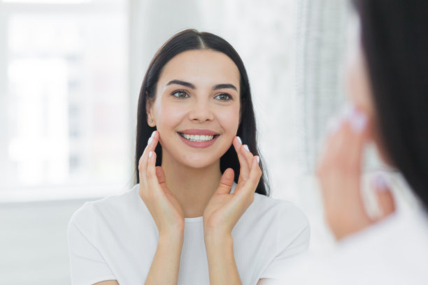 特写照片。一位年轻女子站在家里的镜子前看着自己的倒影。她把面霜涂在脸上，化妆，用手按摩，微笑图片下载