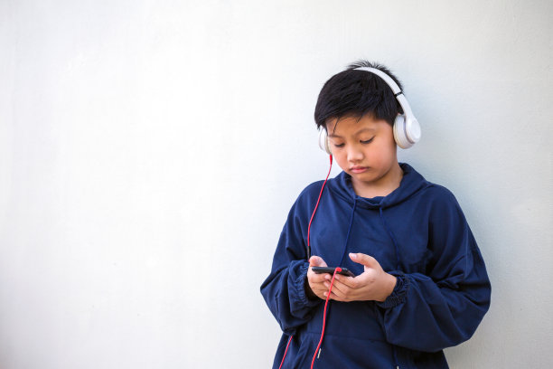 可爱的亚洲男孩，穿着连帽衫，皮肤白皙，在白色的背景下用手机和耳机听音乐。纵向和复制空间图片下载