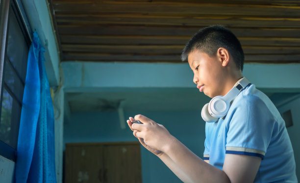 快乐的亚洲男孩坐在卧室里，戴着耳机听音乐，用手机图片下载