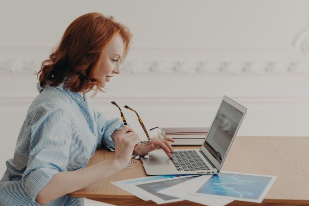 红发自由职业者远程工作，专注于笔记本电脑，坐在放着文件的木桌前，摘下眼镜，上网，看新闻。图片下载