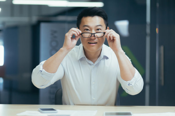 特写照片。一个微笑的亚洲年轻人坐在办公室的桌子前，把眼镜戴在脸上，看着镜头图片下载
