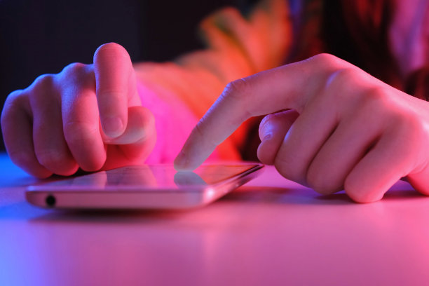 在时尚的霓虹灯下使用手机和笔记本电脑。极富创意的紫外光红蓝鲜艳色彩。女孩的手在黑暗的房间里滚动照片。应用程序在商店销售，社交媒体。图片下载