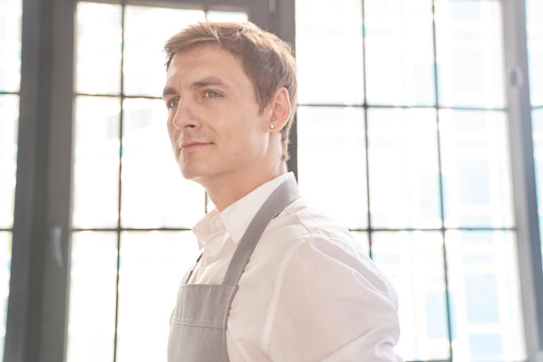 一个穿着灰色围裙的年轻人的特写肖像，若有所思地看着旁边。背景上有一个大窗口，有一个地方可以复制。服务人员。高质量照片图片下载