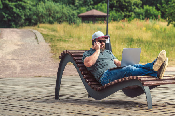 一名交易员拿着笔记本电脑坐在公园的木制躺椅上打电话图片下载