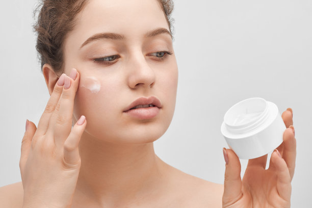 年轻美丽的女孩应用奶油在她的脸，特写。长长的头发，大大的蓝眼睛和清澈的皮肤。图片下载