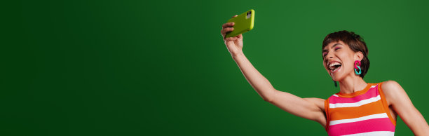 美女用智能手机自拍，站在绿色背景下大笑图片下载