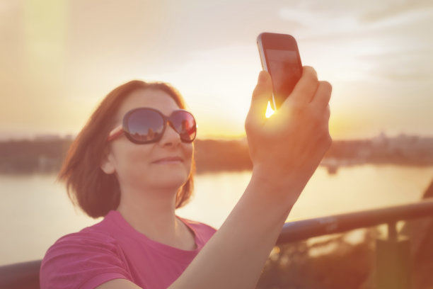 日落时分戴着太阳镜用智能手机自拍的女人图片下载