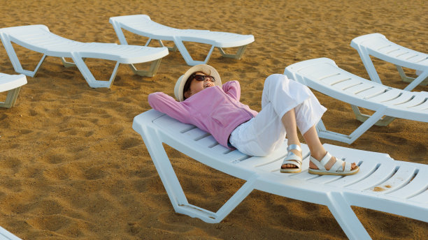 漂亮的小女孩在泳池旁边的太阳躺椅上休息，背景是一排排的太阳躺椅，穿着温暖的衣服。寒冷的天气在夏天的概念图片下载
