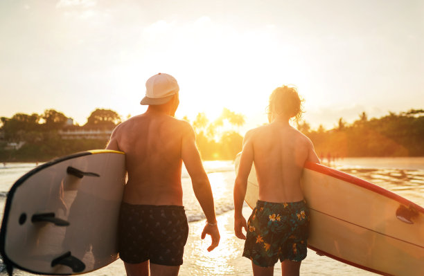 父亲和十几岁的儿子与冲浪板走在沙滩与棕榈树背景下的夕阳。他们微笑着交谈。家庭活动度假概念。图片下载